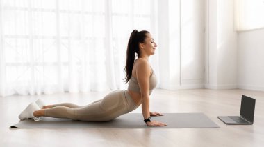 Esnek atletik genç kadın evde yoga yapıyor, kadın fitness minderi üzerinde egzersiz yapıyor, boş ekranlı, yan bakışlı, reklam için kopyalama alanı olan modern bilgisayar üzerinde online ders programı izliyor.