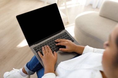 Tanımlanamayan koyu tenli bir kadının siyah ekranlı dizüstü bilgisayar kullanması, reklam için model alması, evdeki kanepede oturan dijital kadın göçebe, bilgisayar klavyesine yazı yazması, yer kopyalaması.