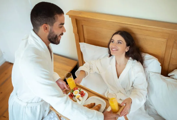 新婚旅行だ ベッドで朝食をとっている幸せなカップル モダンなホテルのベッドルームに座って白いバスローブを着ている妻においしい食べ物をテーブルトレイを与えるロマンチックな驚きを作る男 — ストック写真
