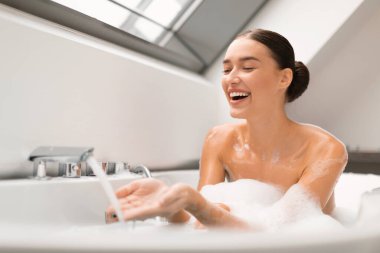 Modern banyodaki banyo küvetinde dinlenirken musluktan akan sıcak suya köpükle banyo yapan mutlu bir kadın. Güzellik rutini ve kaplıca konsepti. Seçici Odaklanma