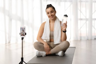 Bayan fitness eğitmeni güzel atletik sporcu genç kadın omuzlarında havluyla yoga minderinde oturur, su şişesini gösterir, su içmeyi tavsiye eder, akıllı telefondan evden yayın yapar.