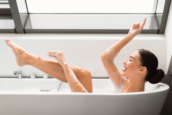安全カミソリで足を剃るハッピーウーマン浴場では 現代の浴室でお風呂に入るデパ地下ルーチンを楽しんでいます 脱毛とボディケア化粧品のコンセプト サイドビューショット — ストック写真