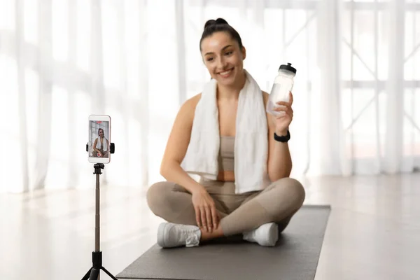 stock image Female fitness instructor beautiful athletic sporty young woman sit on yoga mat with towel on shoulders, showing bottle of water, recommending water drinking, broadcasting from home via smartphone