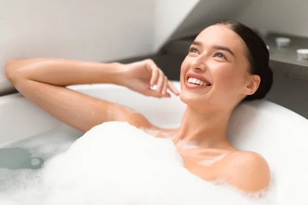 Attractive Female Taking Bath With Foam Enjoying Beauty Ritual In Modern Bathroom Indoors. Happy Woman Lying In White Bathtub Bathing And Relaxing At Home. Wellness And Spa Concept