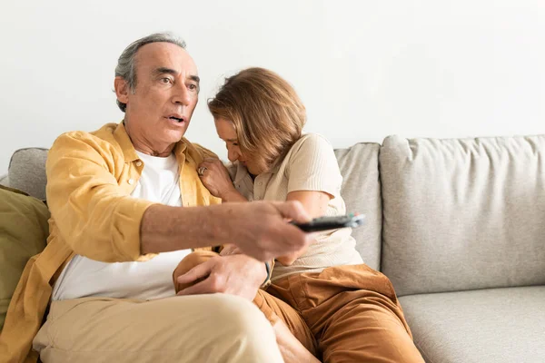 Frightened senior european spouses watching TV together, man holding remote control and woman closing face, sitting on sofa in living room, free space. Scared film and people emotions