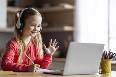 Uzaktan eğitim. Evde dizüstü bilgisayarla video görüşmesi yapan sevimli bir kız kulaklıklı küçük kız Webcam 'e el sallıyor, online ders sırasında öğretmenini selamlıyor, boşluğu kopyalıyor.