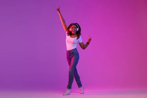 紫のスタジオの背景に明るいネオンライトで音楽やダンスを聞いて無線電話を身に着けているケアフリー若い黒人女性 幸せな正アフリカ系アメリカ人女性お気に入りのプレイリストを楽しんで — ストック写真
