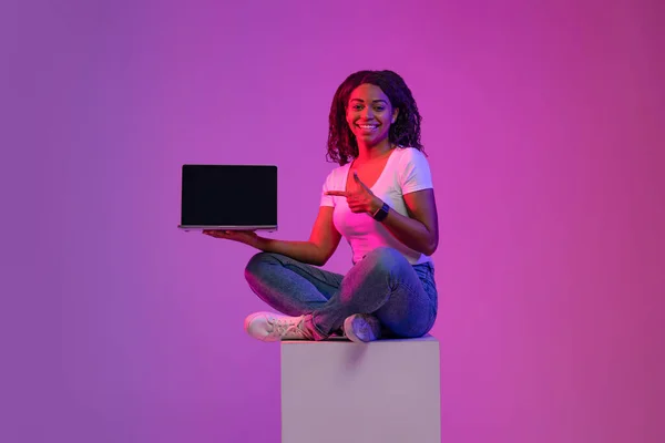 Great Website. Smiling Black Woman Pointing At Blank Laptop Screen While Sitting On Big Cube In Neon Light, Happy African American Lady Showing Free Space For Mockup, Posing Over Purple Background