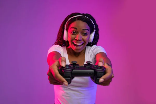 ゲームコンセプト 幸せな興奮アフリカ系アメリカ人女性カメラでジョイスティックを目指して 紫色の背景にネオンライティングに立ちながらゲームパッドで再生ヘッドフォンを身に着けている陽気な黒人女性 — ストック写真