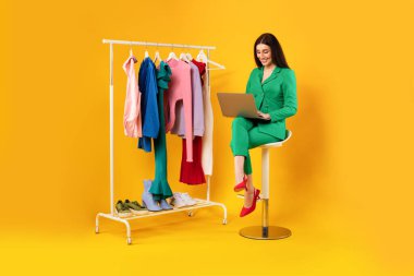 İnternetten alışveriş. Parlak elbiseli, sarı stüdyo arka planlı, uzun boylu bir sandalyede otururken dizüstü bilgisayar kullanan şık bir bayan. Moda ve tarz