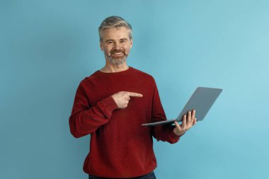 Modern dizüstü bilgisayarı mavi stüdyo arkaplanının üstünde kullanan yakışıklı, orta yaşlı bir adam. Web sörfü yapıyor ya da internette çalışıyor. Bilgisayar ekranına bakıyor, kopyalama uzayı gösteriyor. Modern teknoloji kavramı