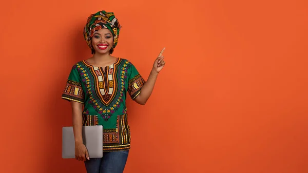 Beautiful smiling attractive millennial african woman wearing national costume freelancer posing with modern pc laptop, ponting at copy space for advert, isolated on orange, banner