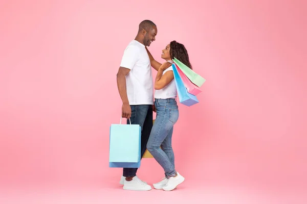 可爱的黑人夫妇拥抱 手持彩色购物袋 站在粉红工作室的背景上 侧视图 消费主义和伟大的销售理念 — 图库照片