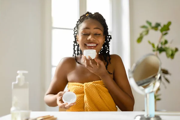 化妆品 护肤概念 快乐的非洲裔美国女人从罐子里抱着奶油 闻到奶油的味道 坐在卧室里 裹着毛巾 自由自在 — 图库照片