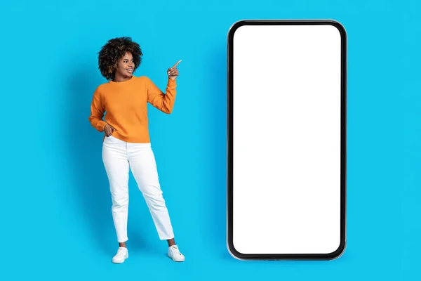 穿着休闲装的迷人的千年黑人女性微笑着 指着蓝色工作室背景的大手机 女性在广告中显示出空白的屏幕复制空间 — 图库照片