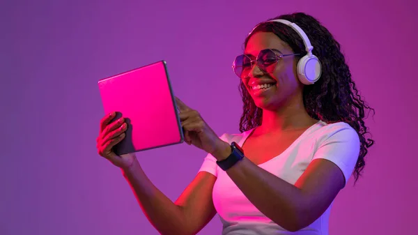 Smiling Young Black Woman In Headphones And Stylish Eyeglasses Using Digital Tablet, Happy African American Woman Browsing Internet On Modern Gadget While Standing In Neon Light On Purple Background