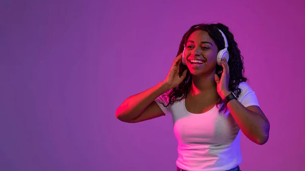 New Playlist. Cheerful Young Black Woman Wearing Wireless Headphones Standing In Neon Light Over Purple Studio Background, Happy African American Female Listening Favorite Music, Copy Space
