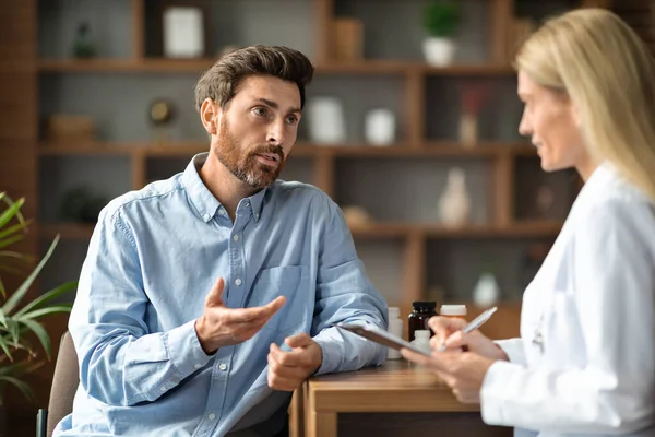Male Patient Sharing Health Problems With Doctor Woman During Meeting In Office, Upset Sick Man Talking To Therapist Lady, Female Physician Holding Clipboard And Making Notes, Selective Focus