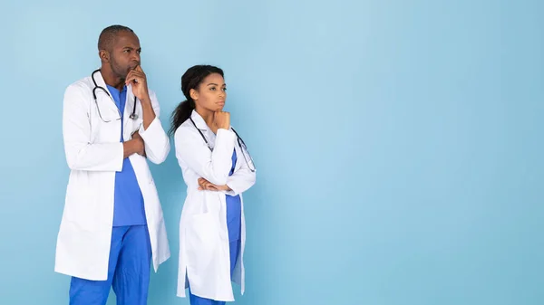 Pensive confident african american medical doctors in white uniform looking at copy space and thinking, standing over blue studio background, panorama