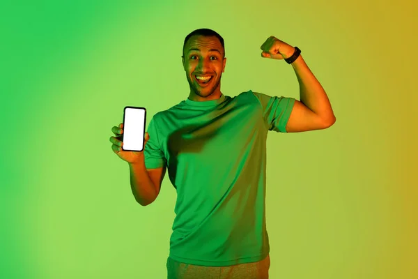 ハッピーブラックスポーツマン広告ワークアウトアプリケーションは ブランクスクリーンと緑のネオンスタジオの背景に立っている彼の足の筋肉で電話を表示します カメラに笑顔 スポーツとガジェット — ストック写真