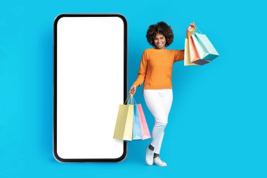 Neşeli, çekici, alımlı genç siyahi bir kadın, beyaz ekranlı büyük bir telefonun başında duruyor ve gülümsüyor, internetten kadın alışverişi yapıyor, mobil uygulama kullanıyor, mavi arka plan, model.