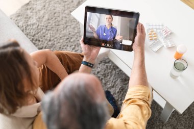 Evdeki e-Sağlık ve teletıp hizmetlerini kullanarak. Üst düzey çift dijital tabletle kanepede oturuyor, online doktorla görüntülü görüşme yapıyorlar ve profesyonel sağlık danışmanlığı alıyorlar.