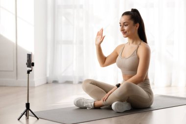 Sanal yoga dersleri konsepti. Yoga eğitmeni. Milenyum güzel, formalı bir kadın. Spor kıyafetleri içinde spor minderinde oturuyor, akıllı telefon kamerasına el sallıyor, öğrencileri selamlıyor, fotokopi çekiyor.