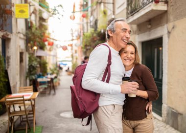 Neşeli yaşlı turistler sırt çantalı, elinde kağıt kahve bardaklarıyla poz veriyorlar Lizbon caddesinde açık kafeli. Emekli Yaşlı Eşler Avrupa 'da Tatil ve Emekliliğin keyfini çıkarıyorlar