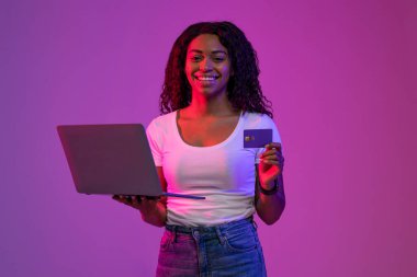 Çevrimiçi ödemeler. Elinde kredi kartı ve laptopuyla gülümseyen Afrikalı Amerikalı kadın mor arka planda neon ışıkta duruyor mutlu siyah kadın internet alışverişi yapıyor ya da fatura ödüyor.