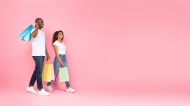 Heyecanlı Afro-Amerikalı çift ellerinde renkli alışveriş torbalarıyla pembe arka planda yürüyorlar ve boş alana, manzaraya bakıyorlar. Alışveriş, perakende, tüketim kavramı