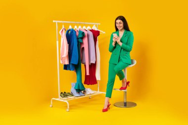 Mutlu moda blogcusu akıllı telefon kullanıyor, giyim kuşağı yanında oturuyor ve sosyal medyayı kontrol ediyor, yazı yazıyor veya çevrimiçi mağazadan alışveriş yapıyor, sarı arka plan