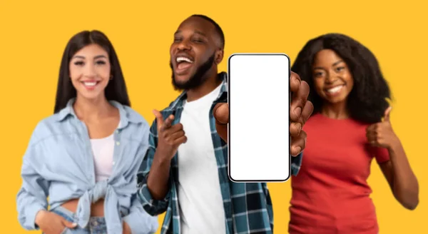 Online ad, offer. Satisfied millennial african american and arab guy and women point finger at phone with blank screen approve website, device and app, isolated on yellow background, studio