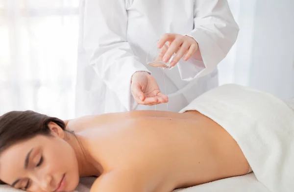 Female therapist applying massage oil on hands before session with young indian woman, relaxed beautiful lady lying on table at spa with closed eyes, enjoying body treatment in wellness center