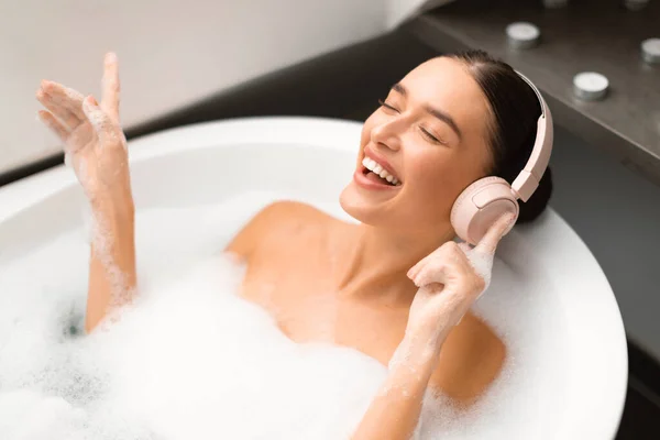 自宅のモダンな浴室にある浴槽に寝そべっているワイヤレスヘッドフォンを身に着けている幸せな女性の入浴と歌 音楽をオンラインで聴いている女性とリラックスしたバスルームの屋内を取ります ウェルネス — ストック写真