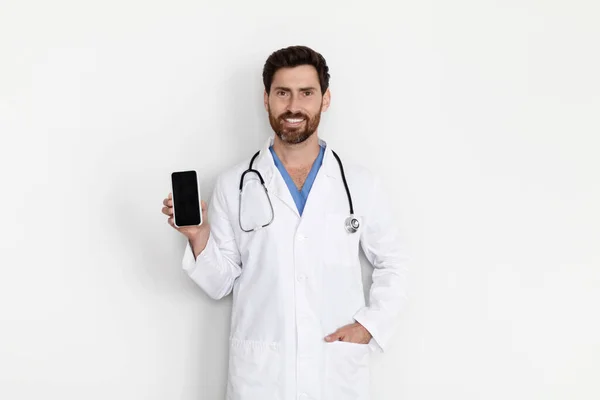 Uśmiechnięty Lekarz Mundurze Demonstrujący Pusty Smartfon Dłoni Stojąc Przy Białej — Zdjęcie stockowe