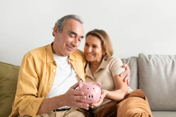 ピンクの貯金箱を手にした幸せなヨーロッパのシニアカップルと抱擁 リビングルームのインテリア フリースペースのソファに座っている 退職貯蓄の概念 — ストック写真