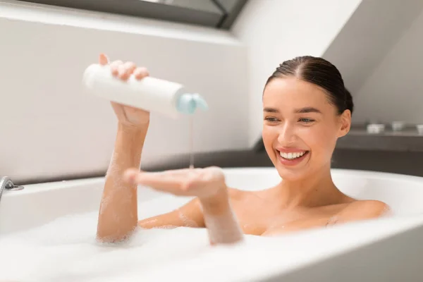 泡でお風呂に入る幸せな女性浴場内の白い浴槽に座ってボトルからシャワージェルを絞ります リラックスして入浴を楽しむ女性に選択的な焦点ルーチン — ストック写真