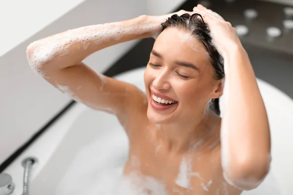 ヘアケア お風呂の中でお風呂に入る白い浴槽に頭を洗うシャンプーを適用する幸せな女性屋内 目を閉じて長い茶色の髪の入浴のための女性のケア 選択的フォーカス — ストック写真