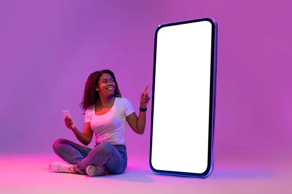 Check This. Happy Black Female Pointing At Big Blank Smartphone In Neon Light, Smiling African American Woman With Cellphone In Hand Advertising New Mobile Application, Collage, Mockup