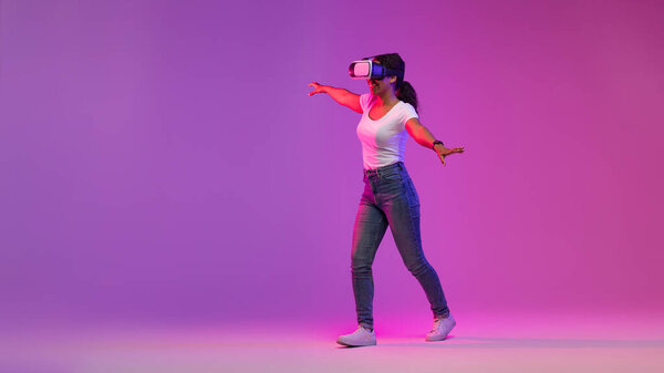 Digital Entertainment. Happy Black Woman In VR Glasses Walking On Purple Background, Excited African American Female Enjoying Virtual Reality Experience, Making Steps In Neon Light, Copy Space