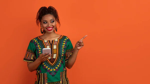 Dating app. Positive smiling beautiful young african american woman in national clothes using smartphone, pointing at copy space for ad, isolated on orange studio background, panorama