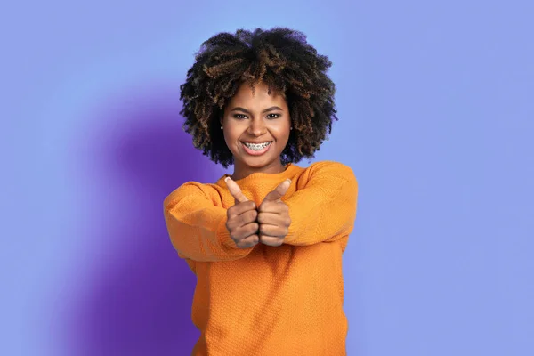 正幸せな陽気な若いですアフリカ系アメリカ人女性でオレンジでブッシーな髪ショー親指アップと笑顔でカメラで紫のスタジオ背景 お勧めします素晴らしい製品やサービス — ストック写真