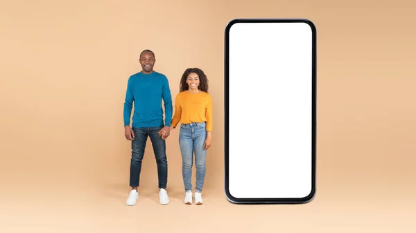 快乐的非洲裔美国配偶站在巨大的手机旁边 在桃子背景 广告申请或网站 全景上都有空白屏幕 — 图库照片