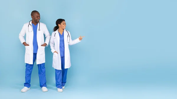 Note it. Positive african american doctors advertising space for advertisement, standing in white medical coats over blue studio background, panorama with free space