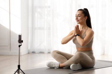 Yoga dersleri online konsept. Çekici, çekici, genç yoga hocasının çevrimiçi dersi var. Spor salonunda antrenman yaparken tripodla yayın yapıyor, namaste jesti yaparak el ele tutuşuyor.