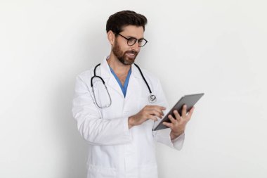 Üniformalı yakışıklı erkek doktorun portresi Dijital Tablet kullanarak, Modern Gadget Danışmanlığı ile gülümseyen doktor hastalar vasıtasıyla çevrimiçi tıbbi sohbet yaparak beyaz arka planda dururken,