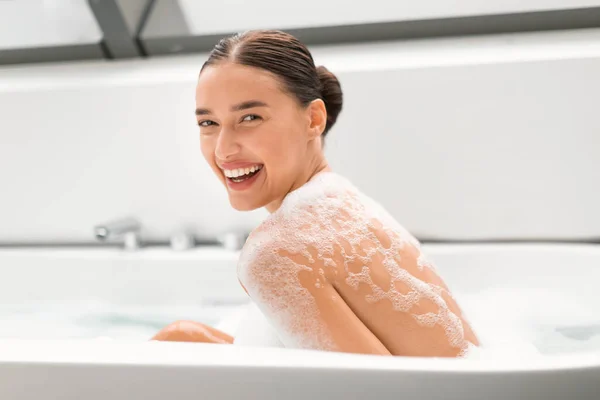 在现代浴室室内 漂亮的女性用泡沫洗澡 微笑着看着相机 洗身体 放松地坐在浴缸里 美丽的礼仪与抚慰的概念 — 图库照片