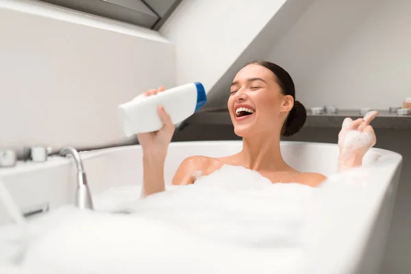 泡でお風呂に入る女の歌声家庭用バスルームのマイクのようなシャワージェルボトル 目を閉じてボディケアルーチンを楽しんでいる女性 選択的フォーカス — ストック写真