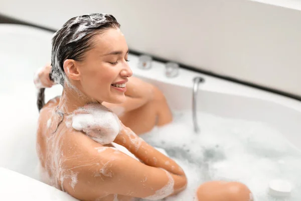 浴室内の浴槽に座っている長い茶色の髪にシャンプーを適用するヘアケアルーチンを楽しんでいる女性の洗髪頭の側面図目を閉じてポーズ ヘアケアの儀式と化粧品の概念 — ストック写真