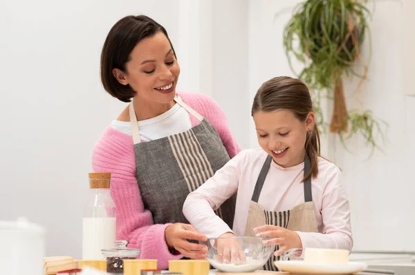 Smiling european millennial lady and small girl in aprons prepare dough for cookies, enjoy lesson in kitchen. Cooking homemade food, hobby in free time at home, mom and daughter relationships